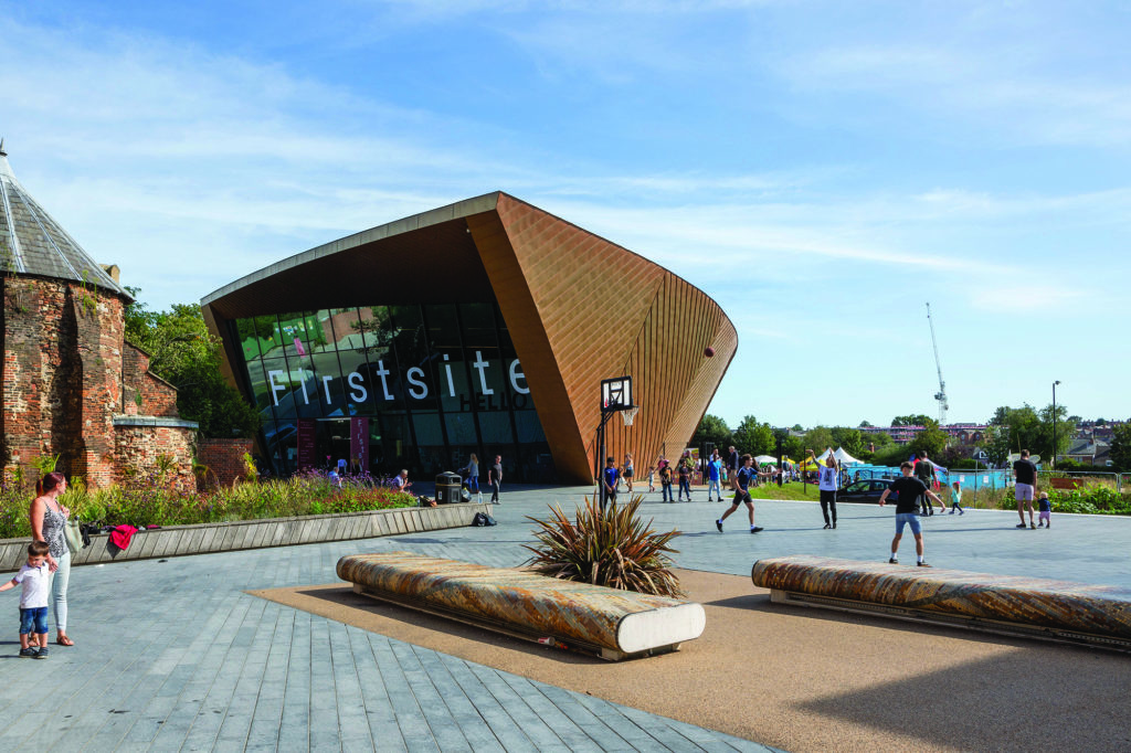 An exterior image of Firstsite museum in Colchester. The building is modern with a glass front saying 'Firstsite'. A group of people are playing a game outside the museum and a woman is walking past with a child.