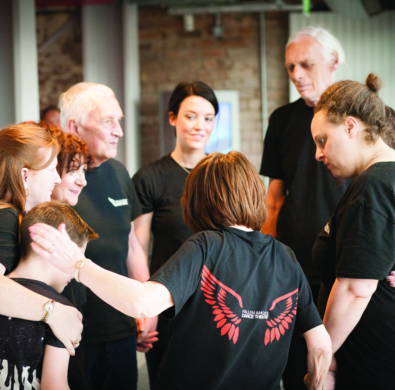 A group of people wearing black t-shirts with a picture of red wings on. They are close together as if hugging in a circle.