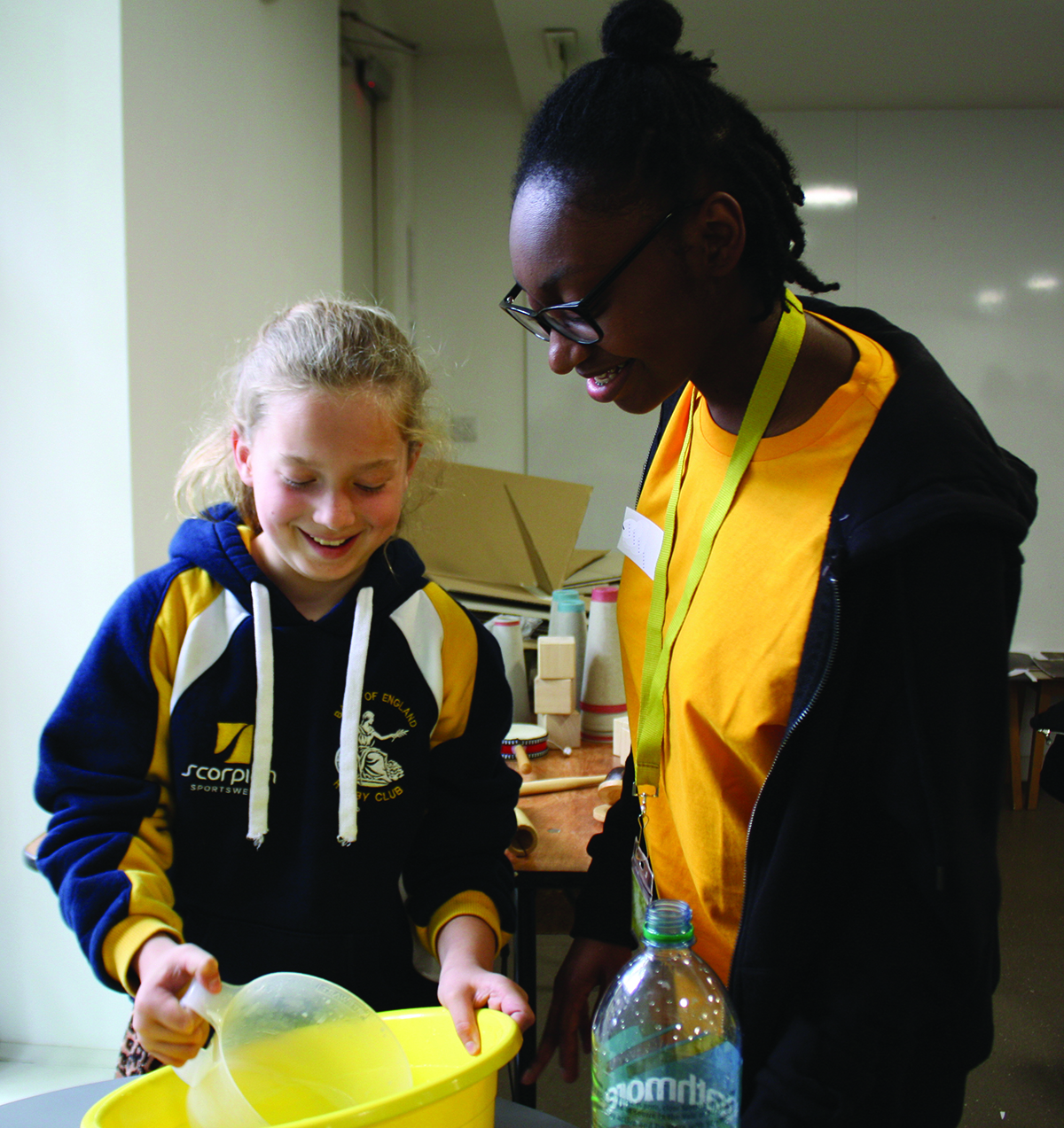 Two girls, one with white skin, one with black skin, are looking together at a bowl and jug. One girl is wearing a blue hoody with yellow and white flashes, the other is wearing a yellow t-shirt with a plain blue hoody.