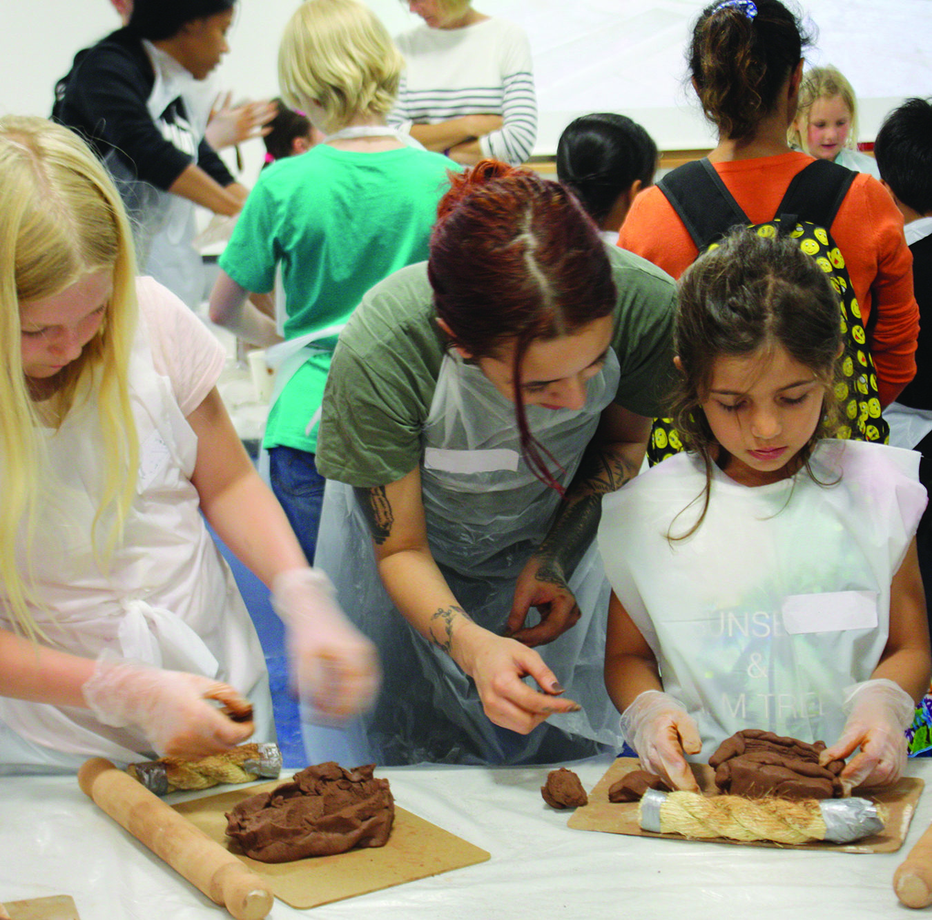A woman is helping two girls to work with clay. All are wearing plastic aprons. In the background more people seem to be engaged in the same activity.