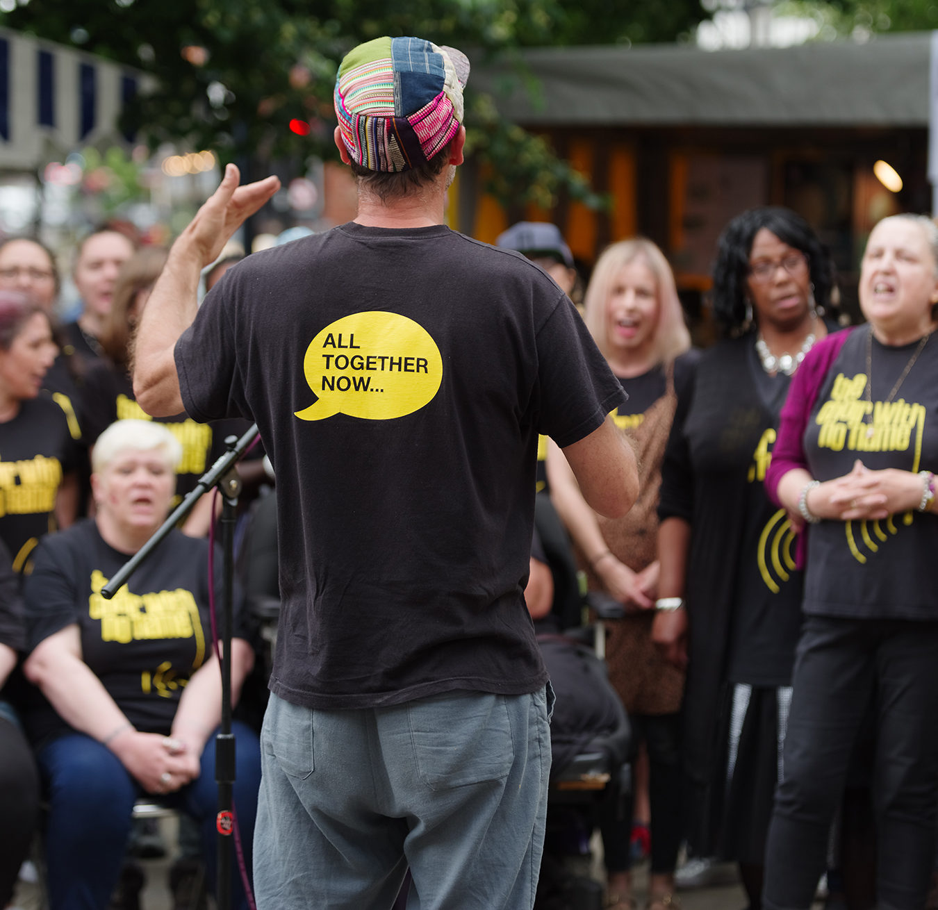 A choir made up of a range of people of different ages and ethnicities. They are wearing t-shirts with The Choir with No Name on. A man is standing in front of them with his back to the camera wearing a black t-shirt with All Together Now on the back.