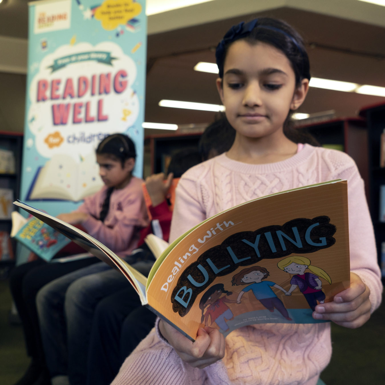A group of children reading in a library. The girl in the foreground has dark hair and a pink jumper and is reading a book called Dealing with Bullying.