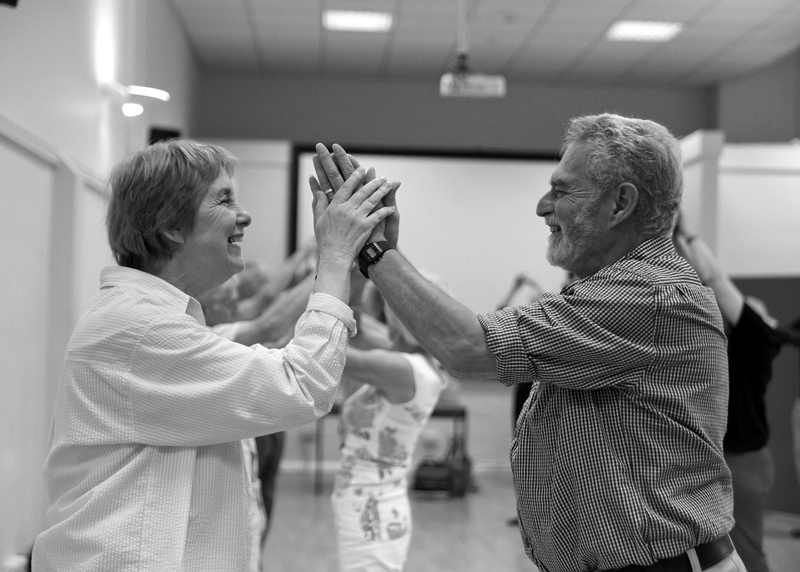 An older couple are facing each other and linking hands. Both are smiling. The woman is wearing a white shirt and the man a checked shirt.