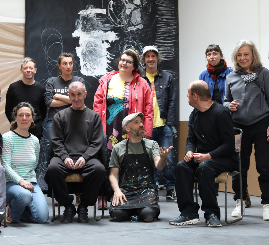 A group of people, some sitting, some standing, in front of an artwork. One man is wearing a dark apron covered with paint splashes.