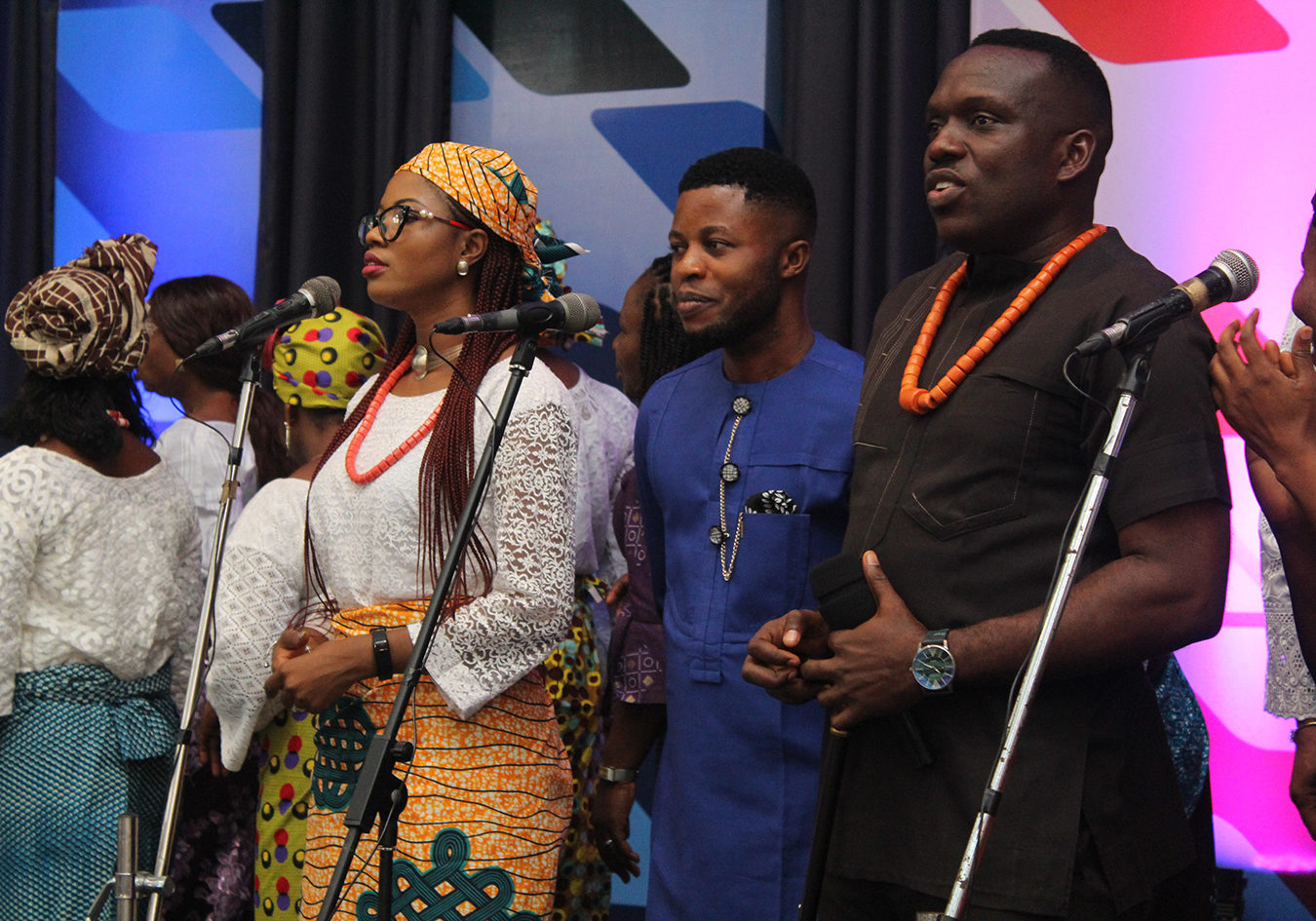 A group of black people in front of microphones. The three people in the foreground are a man wearing black with orange beads, a man wearing blue, and a woman wearing a white top and coloured African print skirt with orange beads and an African print hair wrap.